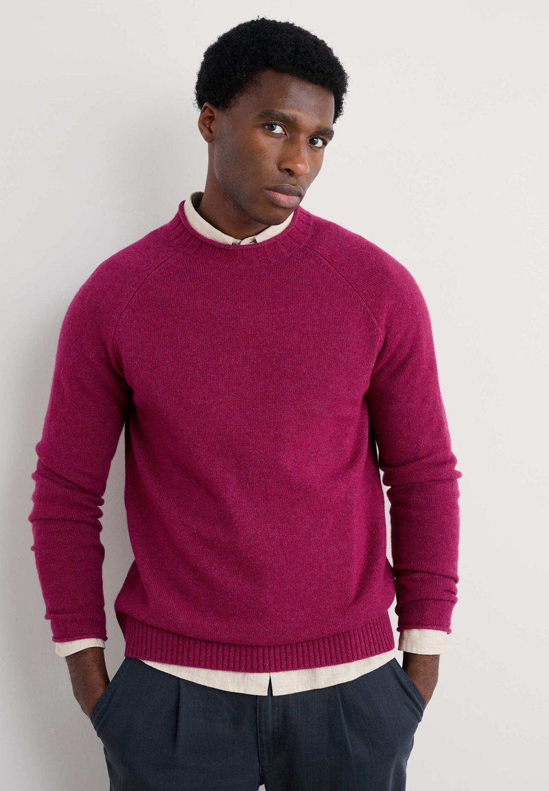 Prigludusi magenta megztinis su apvaliu iškirpimu ir raštuotu apvadu. Yra ilgos rankovės ir dėvimas ant šviesios marškinių su apykakle.