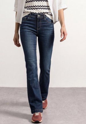 Osoba nosząca ciemnoniebieskie jeansy bootcut, wzorzysty bezrękawnik, białą koszulę i czerwone trampki idąca po szarej podłodze.