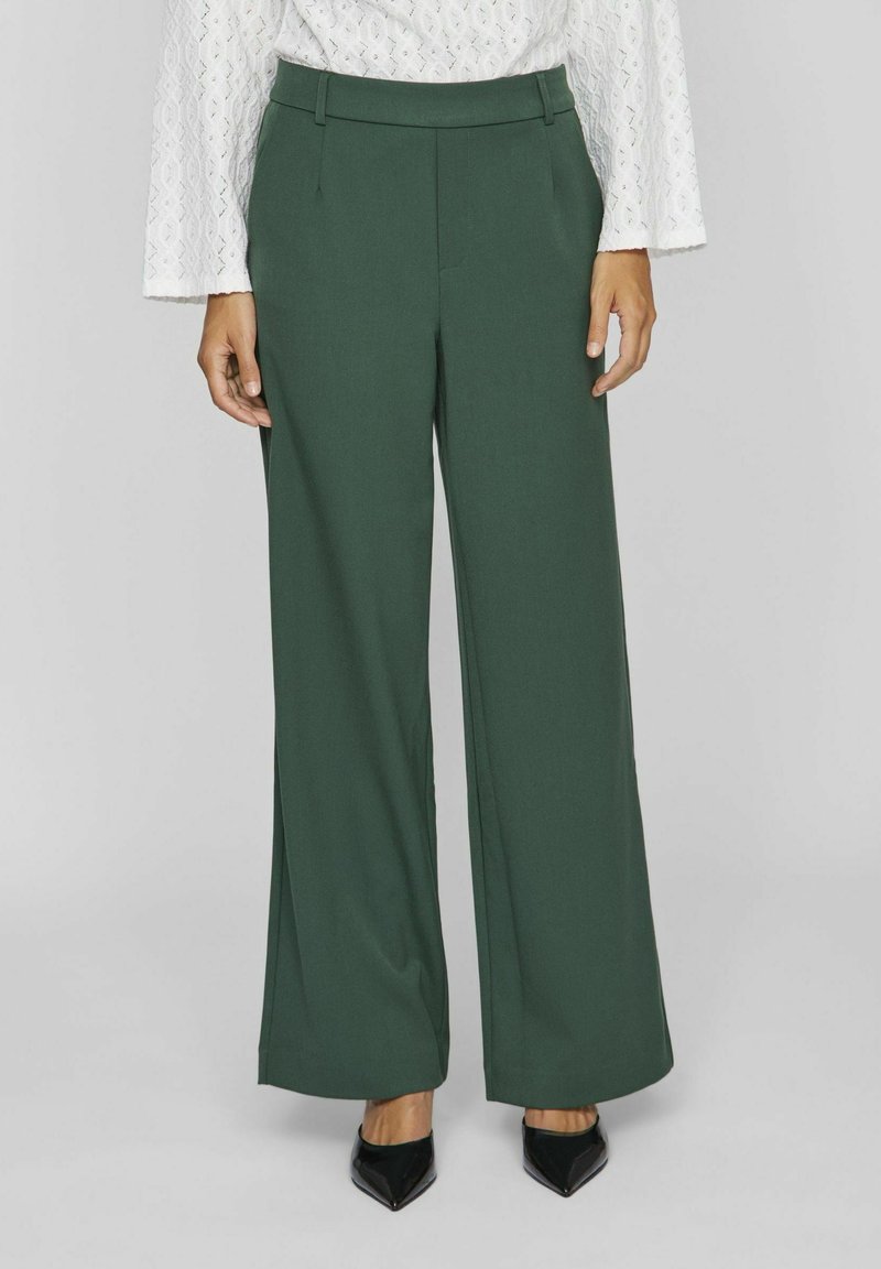 Femme portant un pantalon large vert foncé, un haut en dentelle blanche à manches longues et des talons noirs à bout pointu, debout devant un fond clair.
