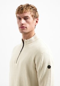 Beige, ribbed knit pullover with a high collar and a black zip. Decorative hexagon patch on the left sleeve adds detail.