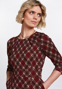 Rotes und schwarzes Karo-Kleid mit drei Viertelärmeln, ausgestattet mit einem runden Halsausschnitt und seitlichen Taschen, aus strukturiertem Stoff gefertigt.