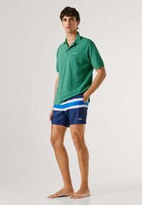 Ung mand, stående barfodet iført grøn polo-shirt og marineblå badeshorts med blå og hvide striber, hænderne i lommerne.