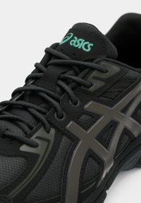 Shoe atlético com um upper de malha preta texturizada, detalhes em cinza, sola de borracha e marcação em verde. Apresenta um sistema de atacadores e um colarinho almofadado.