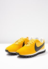 Nike-sneakers med ett ljust gult överlager i mocka, kontrasterande svart swoosh, vit mellansula och texturerad svart gummiyttersula.