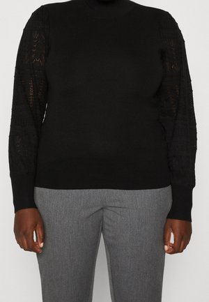 Pull noir à manches longues avec manches en dentelle, associé à un pantalon gris sur mesure, porté par une personne à la peau foncée.