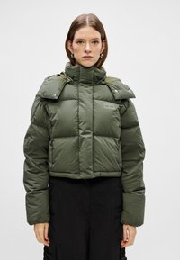 Veste courte matelassée vert foncé avec un col haut, des accents d'épaule à boutons-pression et une texture matelassée. Présente un petit patch logo.