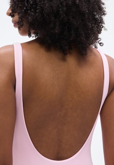 Ροζ μαγιό με σχεδίαση χαμηλής πλάτης, λεπτές τιράντες και ομαλή υφή. Το ύφασμα φαίνεται ελαστικό και εφαρμοστό.