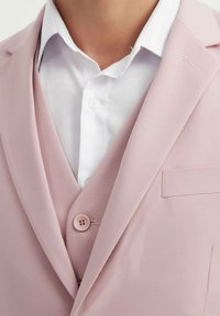 Veste de blazer rose clair avec une finition satinée, revers crantés et une fermeture à un bouton. Positionnée sur une chemise habillée blanche.