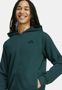 Felpa verde con zip, tessuto strutturato, tasche frontali e dettagli con tre strisce sulle maniche. Presenta un cappuccio con coulisse.