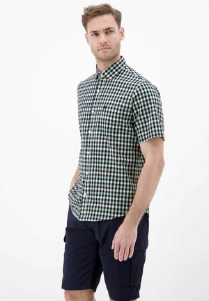 Mężczyzna w zielono-czarnej kraciastej koszuli z krótkim rękawem na guziki oraz granatowych spodenkach cargo, stoi z jedną ręką w kieszeni na białym tle.