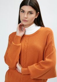 Oranges Sweatshirt mit lockerer Passform, Kabelstrickmuster in der Mitte, gerippte Bündchen und runder Ausschnitt, getragen über einem weißen Rollkragenpullover.