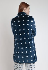 Prenda de forro polar azul marino con un patrón de corazones blancos, que cuenta con cuello y espalda más larga. Textura suave y ajuste relajado, adecuada para ropa cómoda.