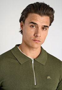 Polo shirt lavorato a maglia verde oliva con zip, dotato di colletto a diaframma e discreto dettaglio del logo sul petto. Texture morbida.