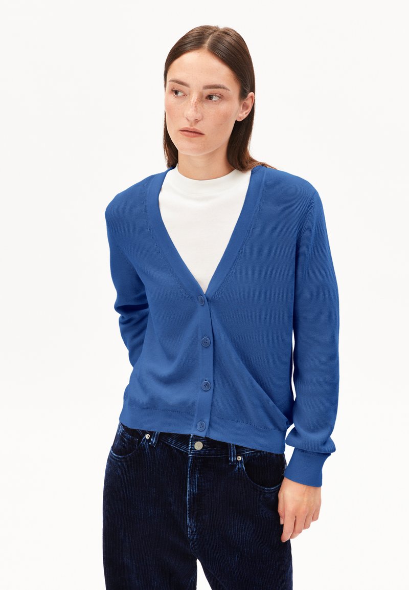 Frau mit glattem braunem Haar, die eine blaue Knopfjacke über einem weißen Oberteil und einer dunkelblauen Cordhose trägt und zur Seite schaut.