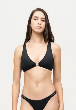 Une femme présente un maillot de bain noir deux pièces avec une fermeture au centre, sur un fond de studio blanc uni.
