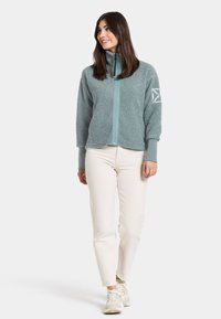 Teal fleecejas met hoge kraag, voorritssluiting en manchetten; heeft een logo op de linkermouw en een zachte, gestructureerde stof.