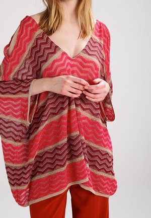 Woman wearing a loose, chevron-patterned red and pink V-neck top with open shoulder cutouts, paired with red pants.