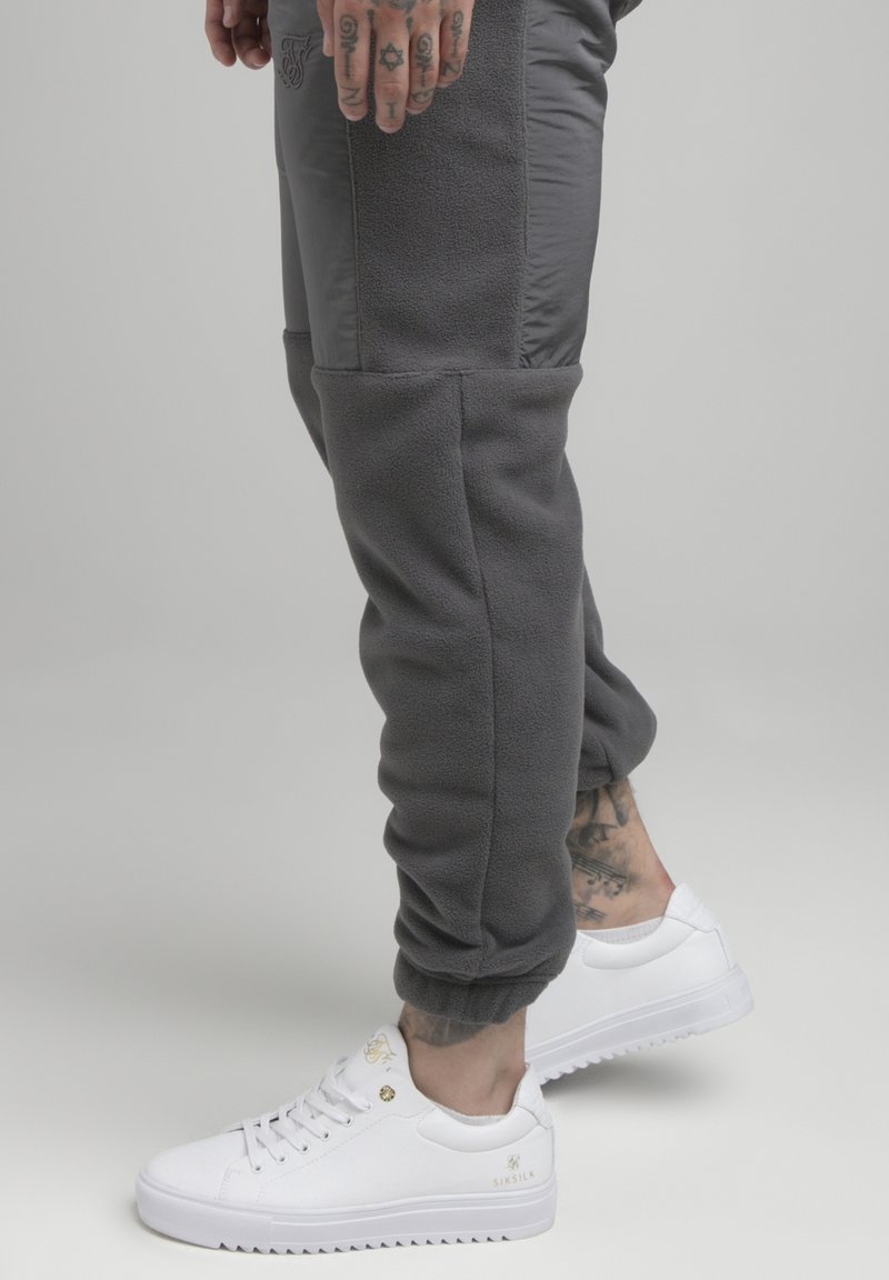Donkergrijze joggers van fleece materiaal met contrasterende gladde panelen, gedragen met witte sneakers met gouden accenten.