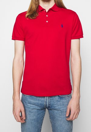 Person trägt rotes kurzärmeliges Polohemd mit drei Knöpfen und kleinem blau besticktem Logo, kombiniert mit blauen Jeans vor einfarbigem Hintergrund.