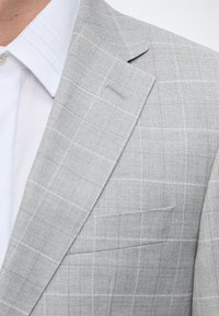 Veste de costume à carreaux gris clair avec un revers étroit portée sur une chemise blanche avec col et bouton visibles.