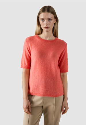 Junge Frau mit glatten blonden Haaren, die einen korallenfarbenen Kurzarmpullover und beigen Hosen trägt, vor einem schlichten hellen Hintergrund stehend.