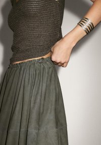 En person justerer snøringen på en grøn plisseret nederdel, iført en tekstureret brun top og flere sølvarmbånd på det ene håndled.