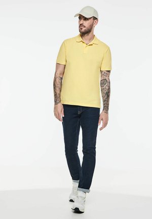 Žluté polo tričko, slim-fit tmavé džíny, bílé tenisky. Béžová čepice. Detaily zahrnují tetování na paži, minimalistický design, hladká textura.