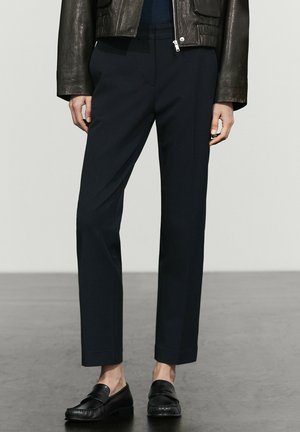 Person trägt schwarze, gerade geschnittene Hose, schwarze Loafers und eine dunkelbraune Lederjacke mit locker an den Seiten hängenden Händen.
