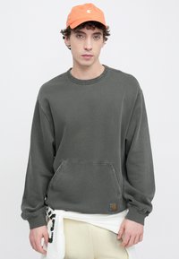 Felpa grigia con scollo rotondo, maniche lunghe e tasca frontale; abbinata a un cappellino arancione con logo; tessuto morbido e casual.