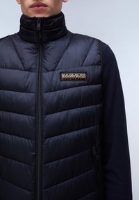 Dunkelblau gesteppter Steppweste mit hohem Kragen, Reißverschluss vorne und Logo-Patch. Glatte Textur, horizontale Nähte, kombiniert mit einer dunkelblauen Fleece-Schicht.