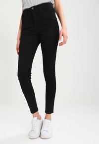 Vaqueros ajustados de talle alto en negro, confeccionados en denim, con cierre de botón, corte slim y diseño hasta los tobillos, combinados con zapatillas blancas.