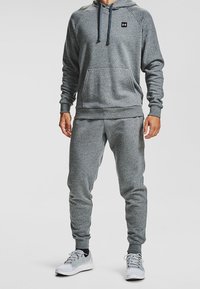 Uomo che indossa una felpa con cappuccio grigia e pantaloni jogger coordinati, con scarpe da ginnastica grigie chiare, in piedi su uno sfondo semplice.