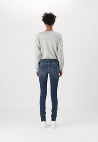 Camisa de manga larga gris claro combinada con jeans ajustados azules. Los jeans tienen un diseño clásico de cinco bolsillos y un ligero estiramiento, terminando por encima de unos zapatos blancos.