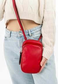Červená kožená crossbody taška s texturovaným povrchem, obdélníkovým tvarem a nastavitelným popruhem. Obsahuje přední kapsu na zip a branding detail.