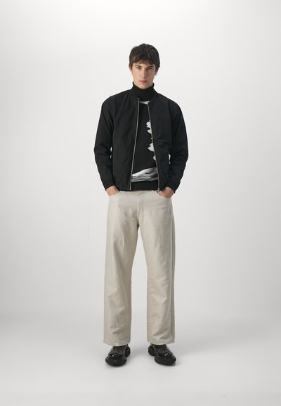 Chaqueta bomber negra con cremallera frontal, camiseta de cuello alto blanca con estampado, pantalones de pierna ancha beige claro y botas chunky negras.