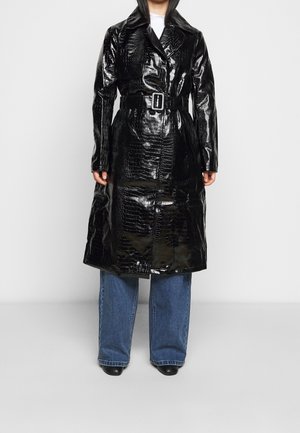 Person iført skinnende sort trenchcoat med krokodillemønster og bælte, over blå jeans og sorte sko, stående mod en ensfarvet baggrund.