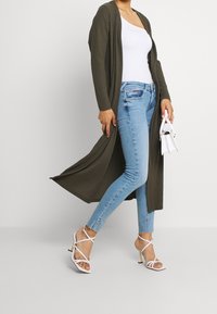 Žena v svetle modrých skinny džínech, bílé topu na jedno rameno, olivově zelené dlouhé cardigan, bílé sandály na podpatku s páskem, držící malou bílou kabelku.