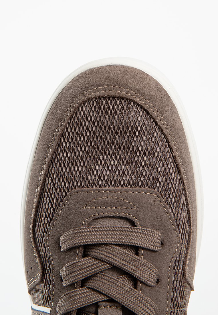 Close-up van de bruine sneakerneus met mesh stof, azen trim, veters en een witte rubberen zool op een witte achtergrond.