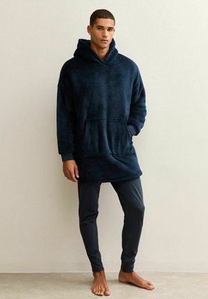 Marineblauwe hoodie-deken sweatshirt, zachte fleece materiaal, voorzak, lange mouwen, gedragen over strakke donkere broeken, blote voeten.