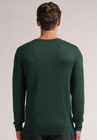 Dunkelgrüner Pullover aus einem weichen Strickmaterial. Verfügt über lange Ärmel, einen runden Hals Ausschnitt sowie Rippenbündchen und Saum.