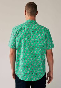 Kurzarmhemd aus gr�ünem Stoff mit einem sich wiederholenden Blumenmuster. Klassischer Kragen und abgerundetem Saum mit subtiler Textur.