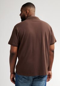 Brązowa polo z kołnierzykiem, wykonana z miękkiej bawełny, z krótkimi rękawami i prostym dołem, noszona z niebieskimi dżinsami.