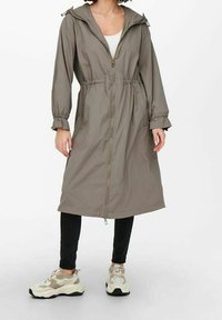 Kvinna klädd i en lång, olivgrön zip-up jacka med cinchad midja, svarta byxor och beige sneakers, stående mot en vit bakgrund.