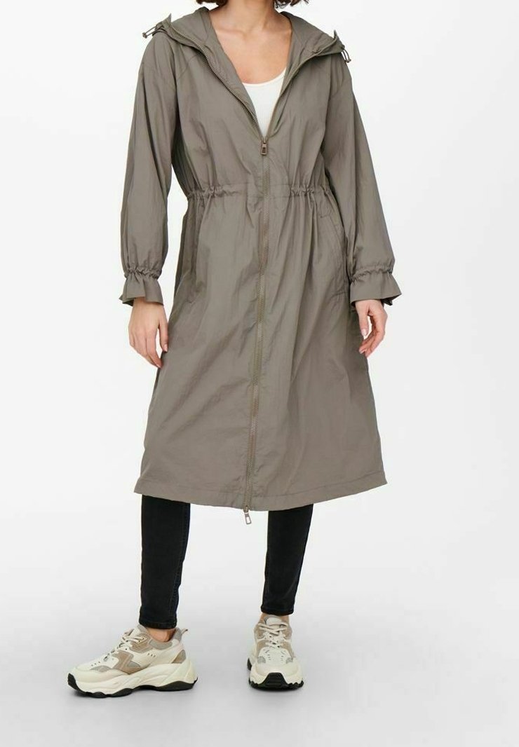 Kvinna klädd i en lång, olivgrön zip-up jacka med cinchad midja, svarta byxor och beige sneakers, stående mot en vit bakgrund.