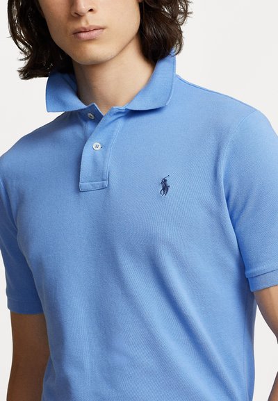 Polo shirt bleu clair avec une finition texturée, patte de boutonnage à trois boutons et logo sombre brodé sur la poitrine gauche. Manches courtes.