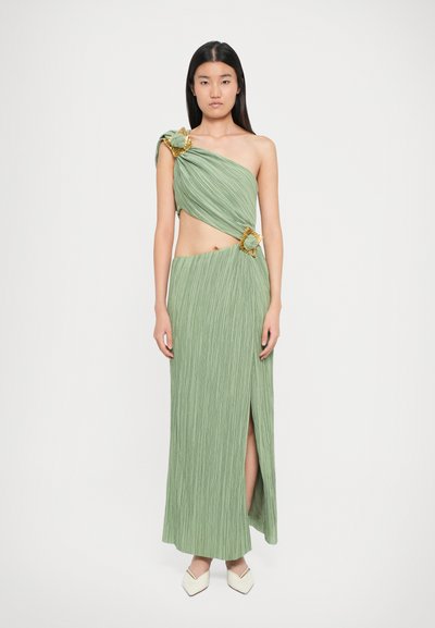 Abito verde a pieghe con spalla unica, caratterizzato da dettagli floreali dorati, uno spacco laterale e una silhouette aderente. Abbinato a scarpe bianche con punta.