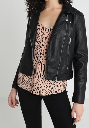 Frau trägt eine schwarze Lederjacke über einer beigefarbenen Bluse mit Tierdruck und schwarzen Hosen, steht vor einem schlichten grauen Hintergrund.