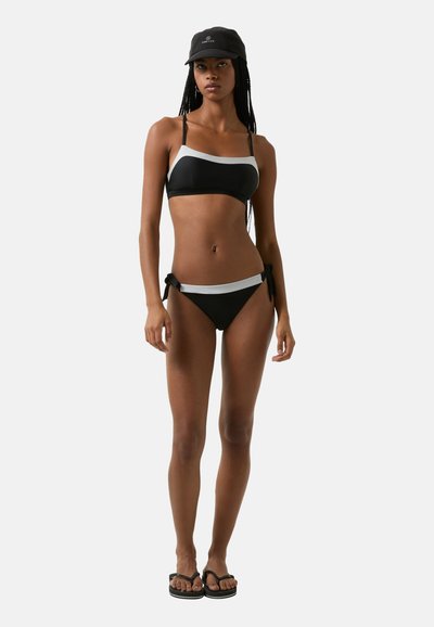 Černé bikiny s bandeau topem a šedými detaily, s bočními stahovacími šňůrkami na kalhotkách. Modelka nosí odpovídající černou čepici a žabky.