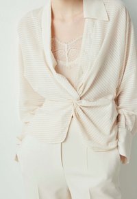Blouse beige clair à boutons avec de fines rayures, coupe décontractée et détail noué à la taille, superposée sur un haut en dentelle.
