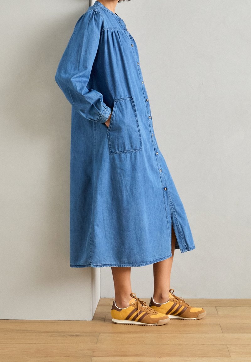 Personne portant une robe en jean bleue mi-longue avec des boutons et des poches à l'avant, associée à des baskets jaunes et marron, debout sur un sol en bois.